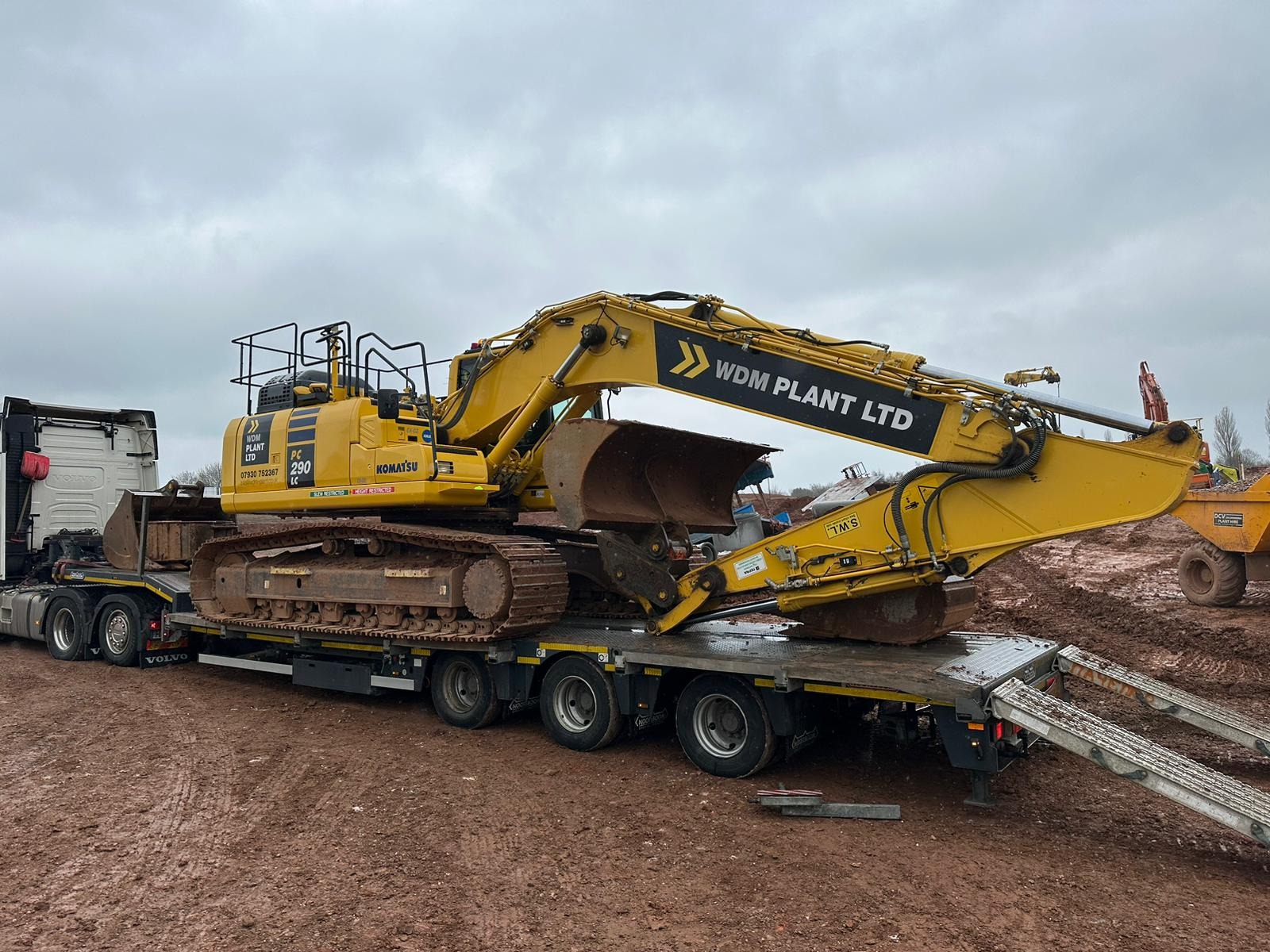 Żółta koparka Komatsu PC290LC z logo WDM Plant Ltd transportowana na niskopodwoziowej lawecie Volvo w błotnistym terenie, pochmurne niebo w tle.
