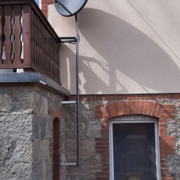 Mont-Sat - Czarna antena satelitarna zamontowana na metalowym stelażu przy balkonie budynku z kamienną elewacją i ceglanymi elementami dekoracyjnymi wokół okien.