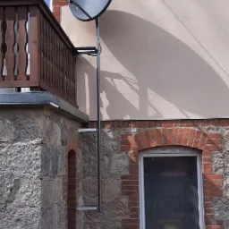 Czarna antena satelitarna zamontowana na metalowym stelażu przy balkonie budynku z kamienną elewacją i ceglanymi elementami dekoracyjnymi wokół okien.