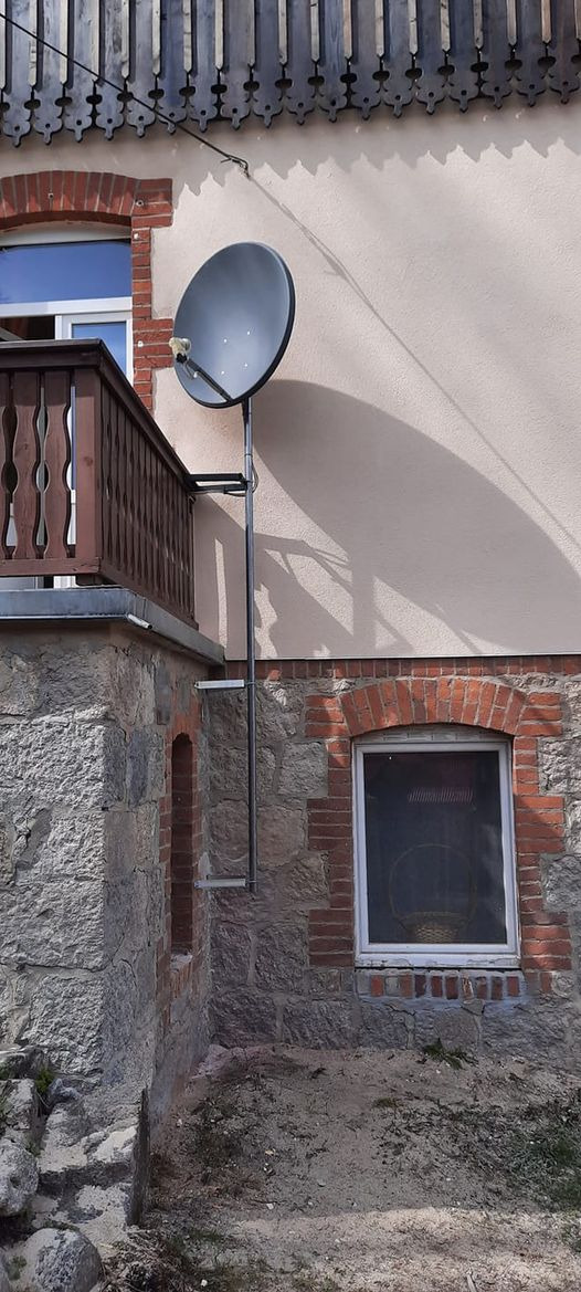 Czarna antena satelitarna zamontowana na metalowym stelażu przy balkonie budynku z kamienną elewacją i ceglanymi elementami dekoracyjnymi wokół okien.