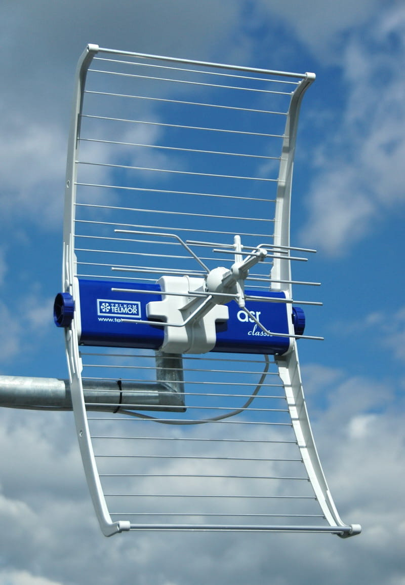 Biała antena typu Yagi-Uda z niebieskim elementem centralnym, zamontowana na metalowym maszcie, na tle błękitnego nieba z białymi obłokami.