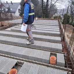 KONSPRO SPÓŁKA Z OGRANICZONĄ ODPOWIEDZIALNOŚCIĄ - Budowa stropu Teriva: inżynier budowy z dokumentami kontroluje ułożenie pustaków szalunkowych i zbrojenia na budowie domu jednorodzinnego, w tle widoczny przenośny WC.