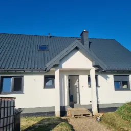 Nowoczesny budynek mieszkalny jednorodzinny z ciemnoszarą dachówką, białymi ścianami i szarymi ramami okiennymi, widziany w słoneczny dzień z niebieskim niebem w tle. Na pierwszym planie widoczny...