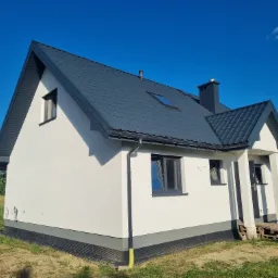 Nowoczesny, biały dom jednorodzinny z ciemnoszarą dachówką i stolarką okienną, widok z zewnątrz na tle błękitnego nieba, widać fragment ogrodu i palety przed wejściem.