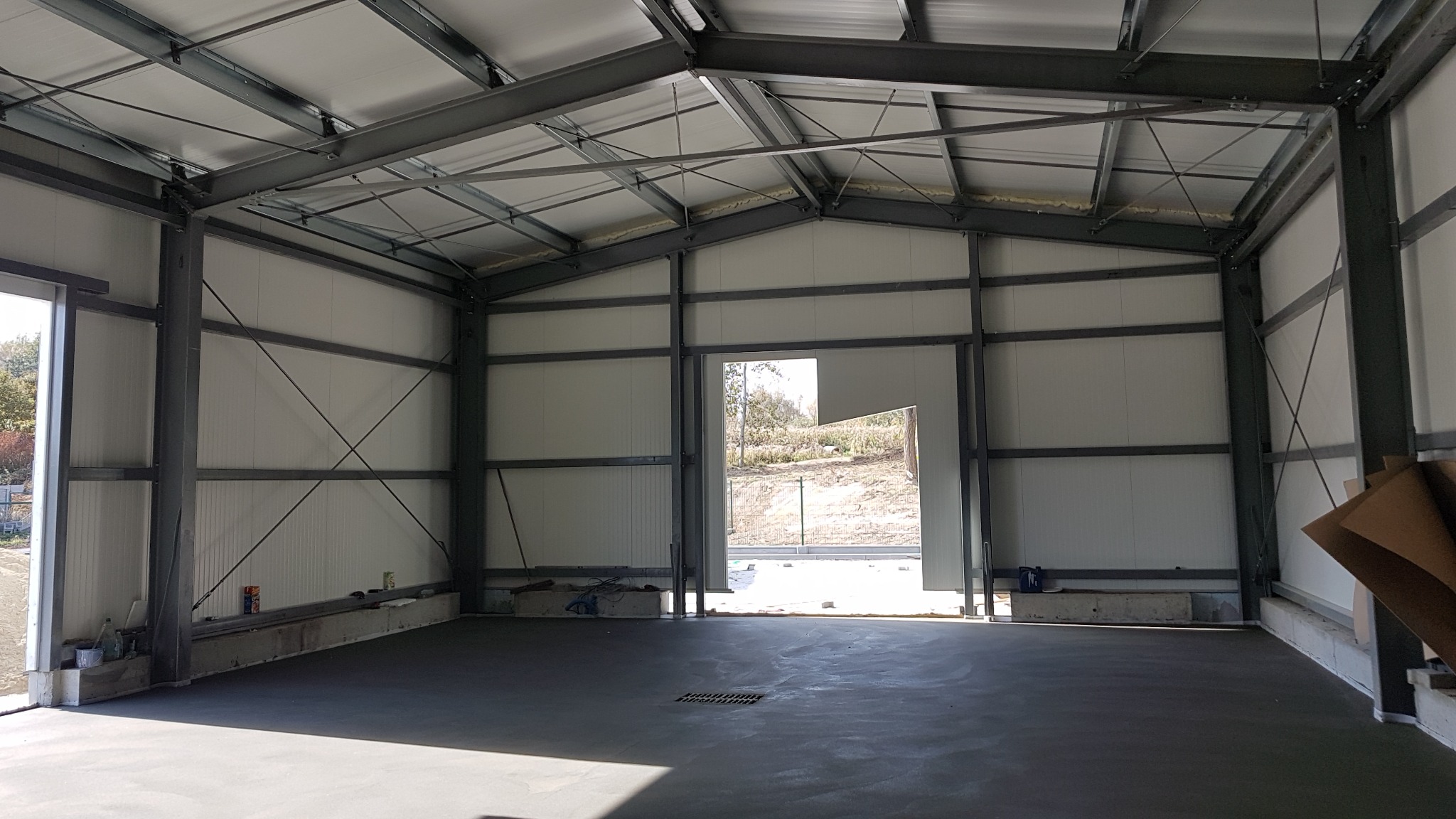 Wnętrze hali w konstrukcji stalowej z wylewką betonową, widoczne elementy izolacji na dachu, ściany z paneli warstwowych, na posadzce kratka ściekowa.