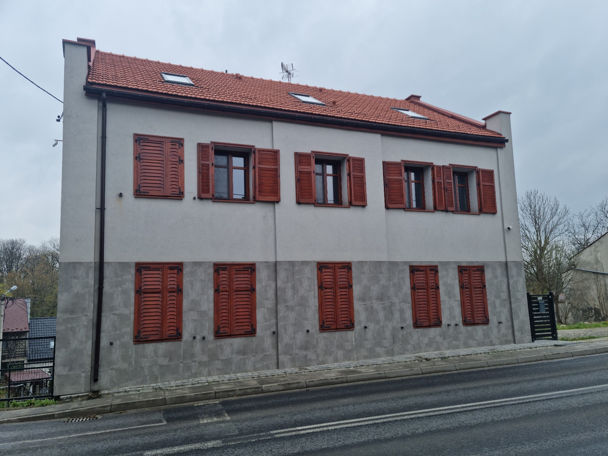 Dwukondygnacyjny budynek z szarą elewacją, czerwonym dachem i brązowymi okiennicami, widoczny z poziomu ulicy.