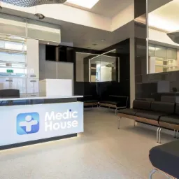 Nowoczesne wnętrze recepcji Medic House: biała lada z logo, czarne sofy, lustrzane ściany i designerskie lampy sufitowe. Minimalistyczny styl, jasne oświetlenie.
