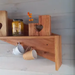 Drewniana półka narożna z dekoracjami: lampiony z wyciętymi sercami, słoik z przetworami i wiszące filiżanki na tle białej boazerii.