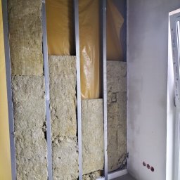 Daw-Bud Dawid Bobola - Wnętrze w trakcie prac: widoczne metalowe profile, wełna mineralna jako izolacja, folia paroizolacyjna oraz fragment otynkowanej ściany z gniazdkami elektrycznymi.