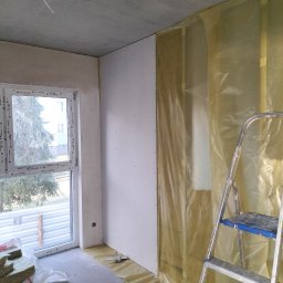 Daw-Bud Dawid Bobola - Wnętrze w trakcie prac wykończeniowych: ściana pokryta płytami gipsowo-kartonowymi z widocznymi wkrętami, zabezpieczona folią ochronną, drabina aluminiowa, okno z widokiem na drzewo i ocieplenie...