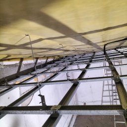 Daw-Bud Dawid Bobola - Sufit podwieszany w trakcie montażu: widoczne metalowe profile, elementy mocujące i częściowo ułożona warstwa izolacyjna w odcieniach żółci.
