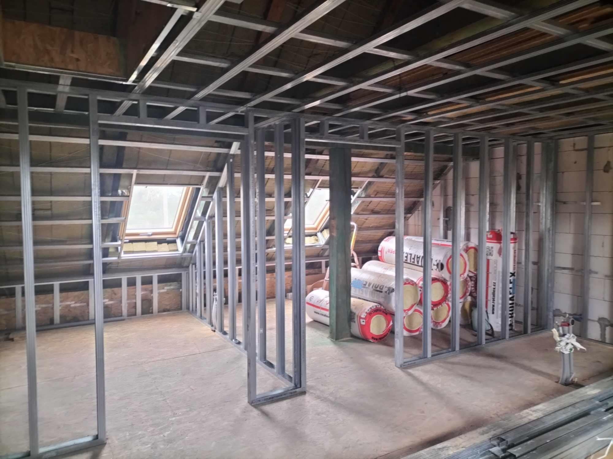 Konstrukcja szkieletowa z metalowych profili w trakcie montażu na poddaszu, widoczne okna dachowe i materiały izolacyjne w rolkach.