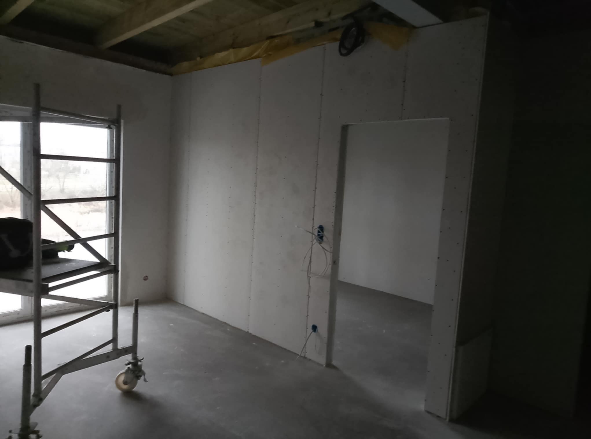 Wnętrze w trakcie prac wykończeniowych: ściany pokryte płytami gipsowo-kartonowymi, widoczne puszki elektryczne z wystającymi przewodami, metalowy ruszt przy oknie, surowy beton na podłodze...
