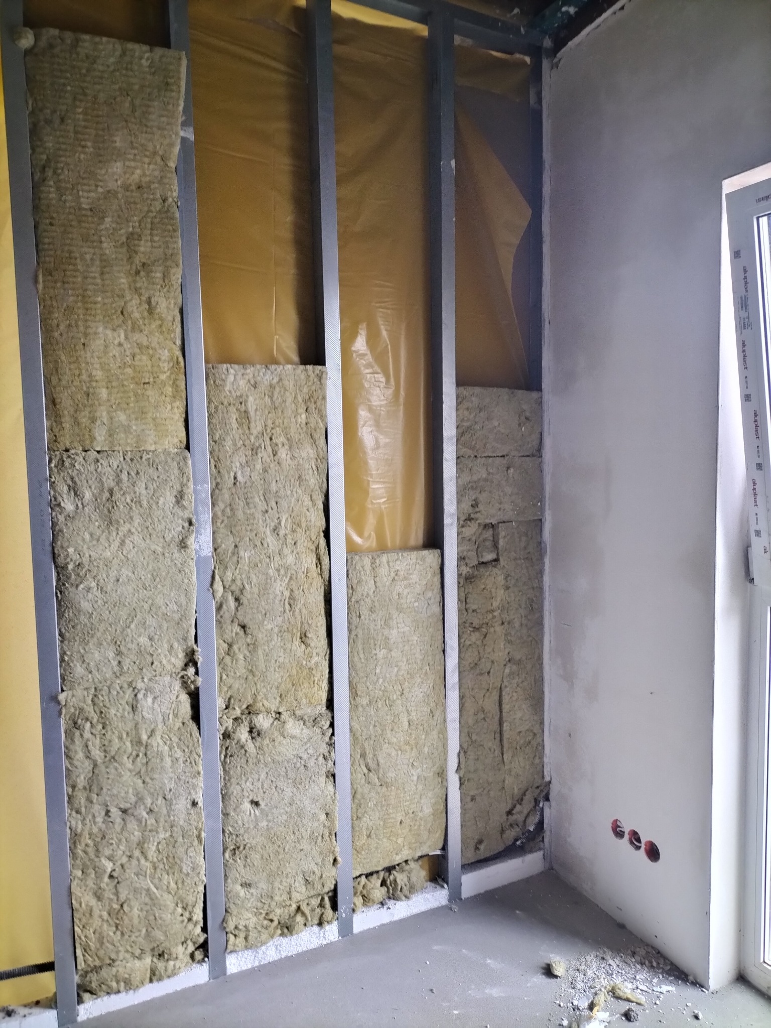 Wnętrze w trakcie prac: widoczne metalowe profile, wełna mineralna jako izolacja, folia paroizolacyjna oraz fragment otynkowanej ściany z gniazdkami elektrycznymi.