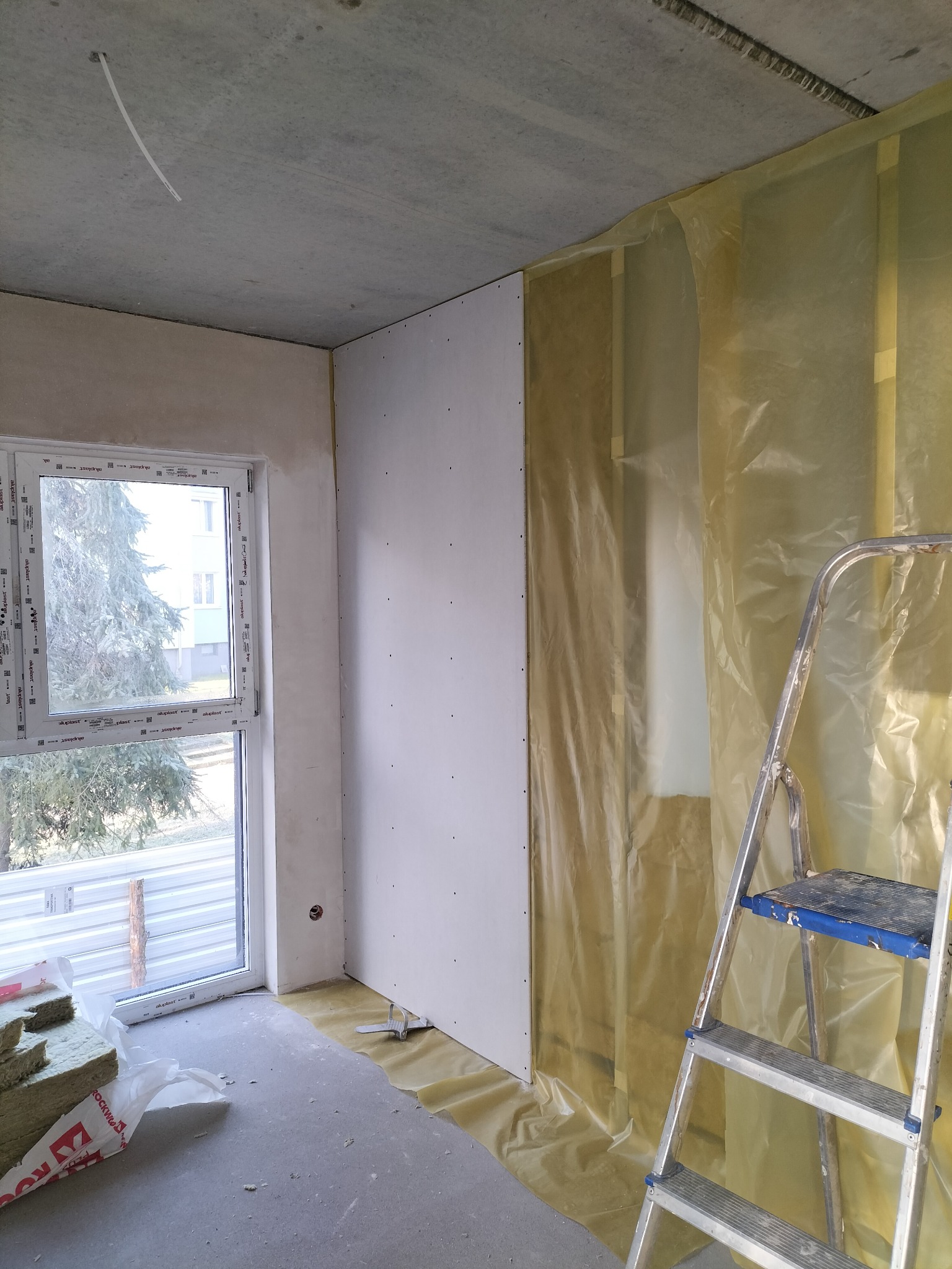Wnętrze w trakcie prac wykończeniowych: ściana pokryta płytami gipsowo-kartonowymi z widocznymi wkrętami, zabezpieczona folią ochronną, drabina aluminiowa, okno z widokiem na drzewo i ocieplenie...