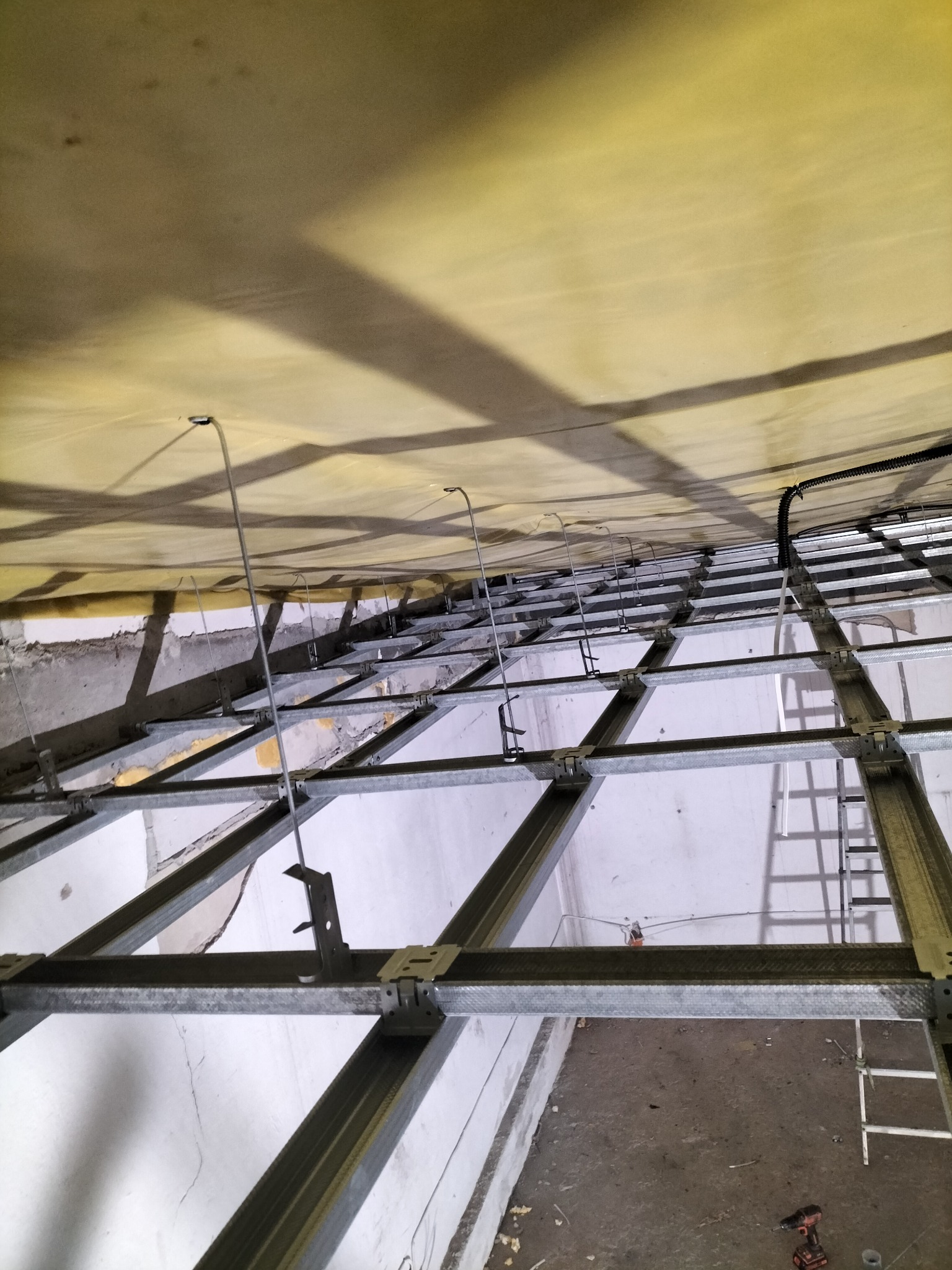 Sufit podwieszany w trakcie montażu: widoczne metalowe profile, elementy mocujące i częściowo ułożona warstwa izolacyjna w odcieniach żółci.