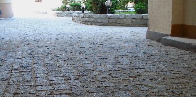 Ułożona szara kostka granitowa tworząca ścieżkę, z widocznym fragmentem budynku z prawej strony i niskim murem z kamienia z roślinami i lampami w tle.