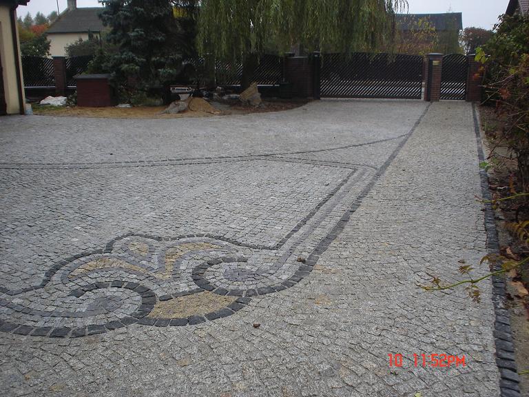 Podjazd z szarej kostki granitowej z dekoracyjnym wzorem w odcieniach szarości i beżu, obramowany czarną kostką, prowadzący do bramy wjazdowej z ceglanymi słupkami.