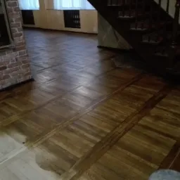 Fragment parkietu w trakcie renowacji, część podłogi wycyklinowana i pokryta lakierem, widoczne schody i ceglana ściana z lustrem w tle.