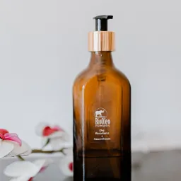 Butelka z brązowego szkła z pompką, etykietą BioOleo Cosmetics, olejem Macadamia i napisem Sweet Dream, obok gałązka z białymi kwiatami.