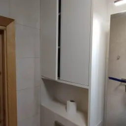 Biała szafka łazienkowa na wymiar z częściowo otwartymi drzwiami, wbudowana nad toaletą, z rolką papieru toaletowego na półce i chromowanym przyciskiem spłuczki.