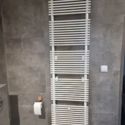 Biały, drabinkowy grzejnik łazienkowy na szarej, płytkowej ścianie, widoczny fragment toalety i uchwyt na papier toaletowy