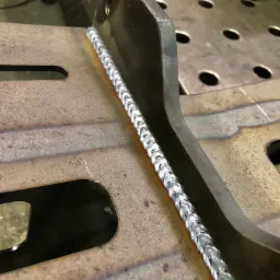 Świeżo wykonany spaw na metalowej konstrukcji, widoczne charakterystyczne łuski spoiny na tle stołu spawalniczego z otworami.