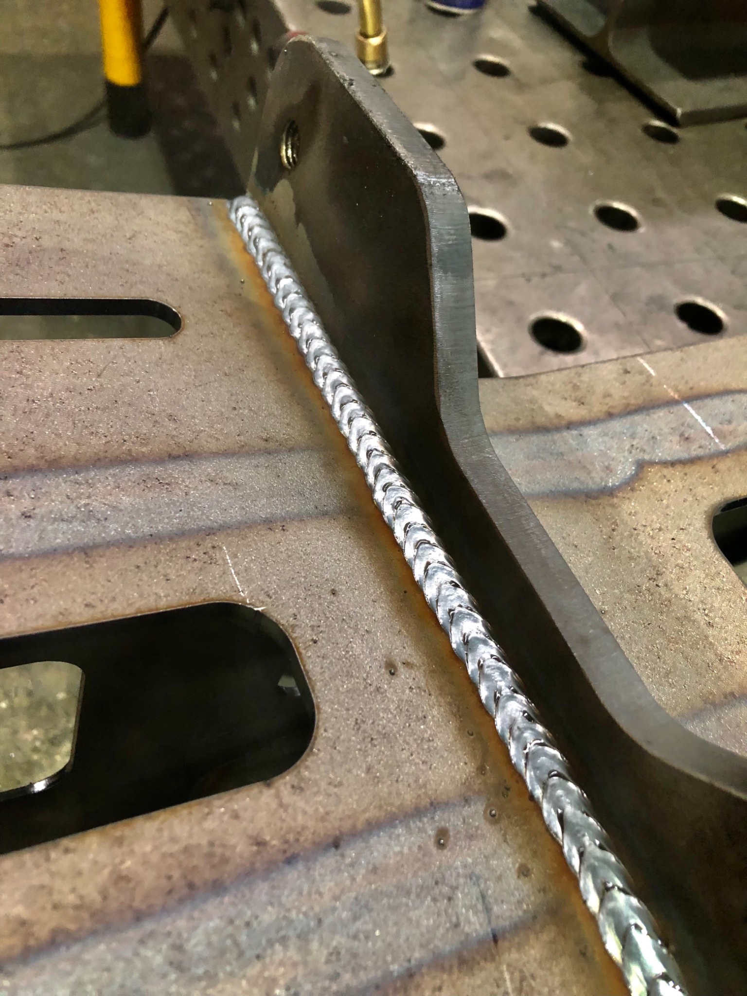 Świeżo wykonany spaw na metalowej konstrukcji, widoczne charakterystyczne łuski spoiny na tle stołu spawalniczego z otworami.