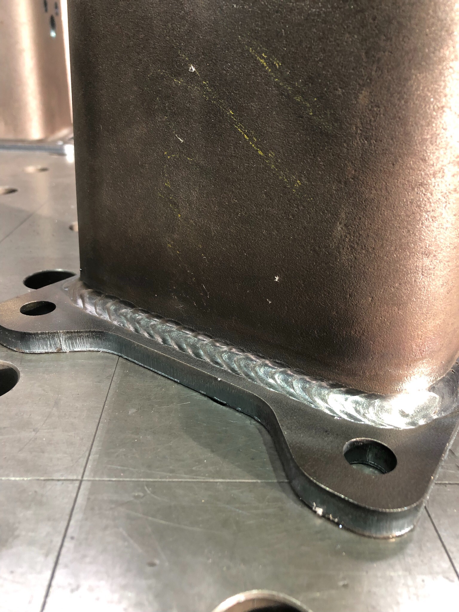 Metalowy element konstrukcyjny z widocznym, świeżym spawem, leżący na perforowanym stole spawalniczym.