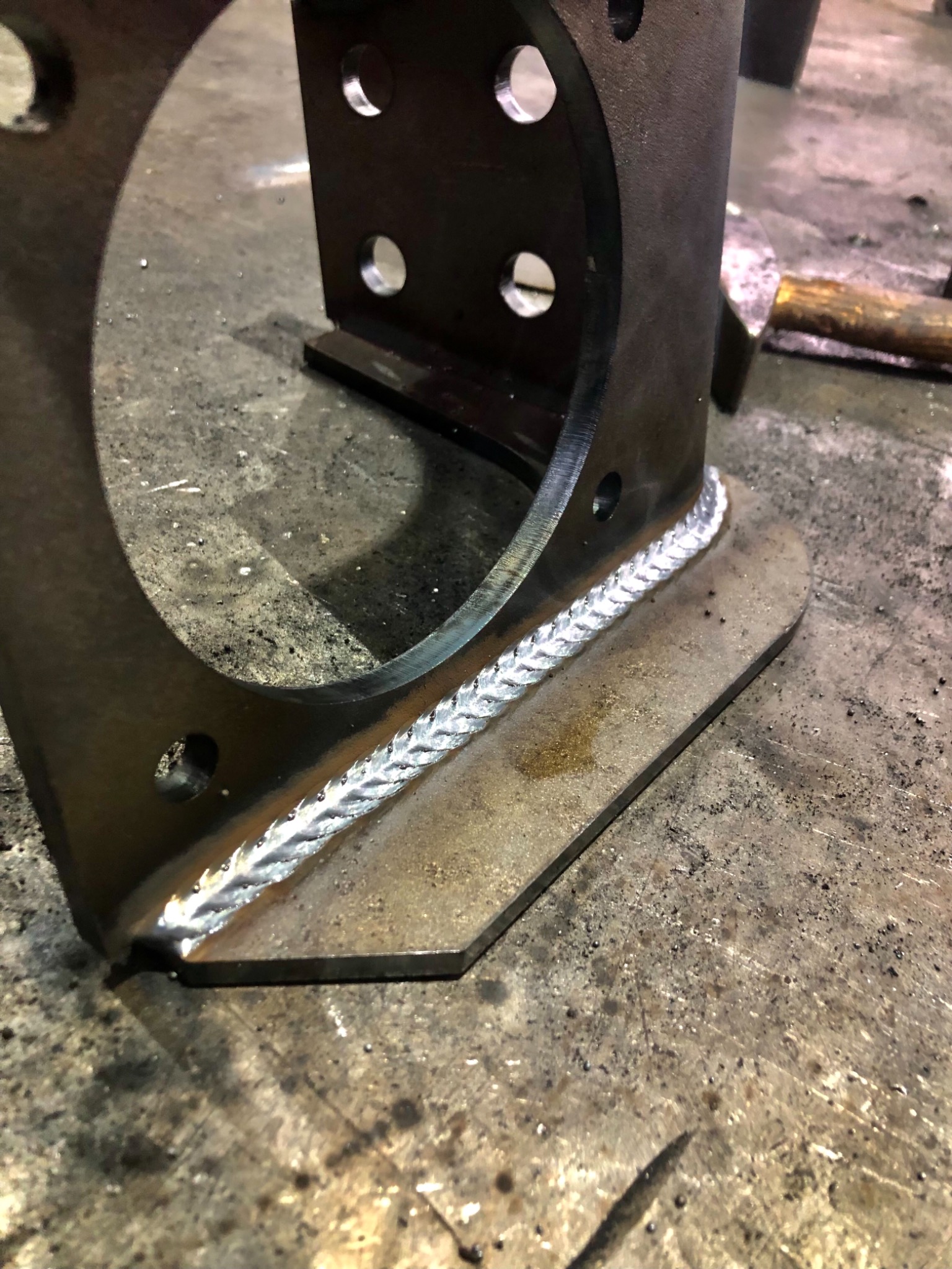 Metalowa konstrukcja spawana z widocznym szwem spawalniczym o strukturze łusek, z otworem okrągłym i rzędem mniejszych otworów, leżąca na brudnym metalowym stole.