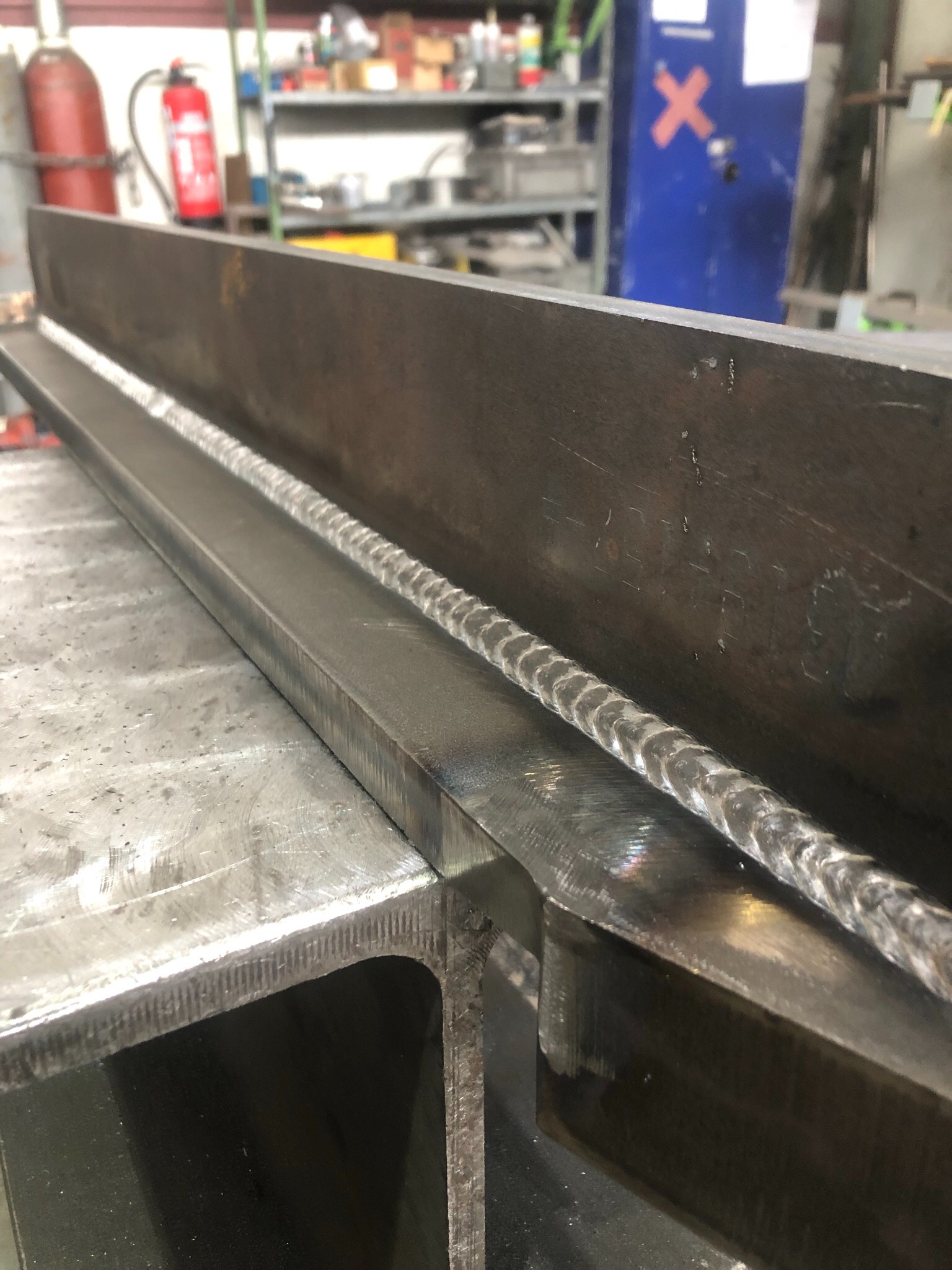 Spoina pachwinowa na elemencie stalowym, widoczny regularny wzór spawu, tło warsztatowe z butlą gazową i szafką.