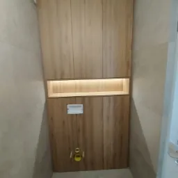 Wnęka z drewnianymi szafkami w łazience, z podświetloną półką i zabudową toalety w odcieniach drewna.