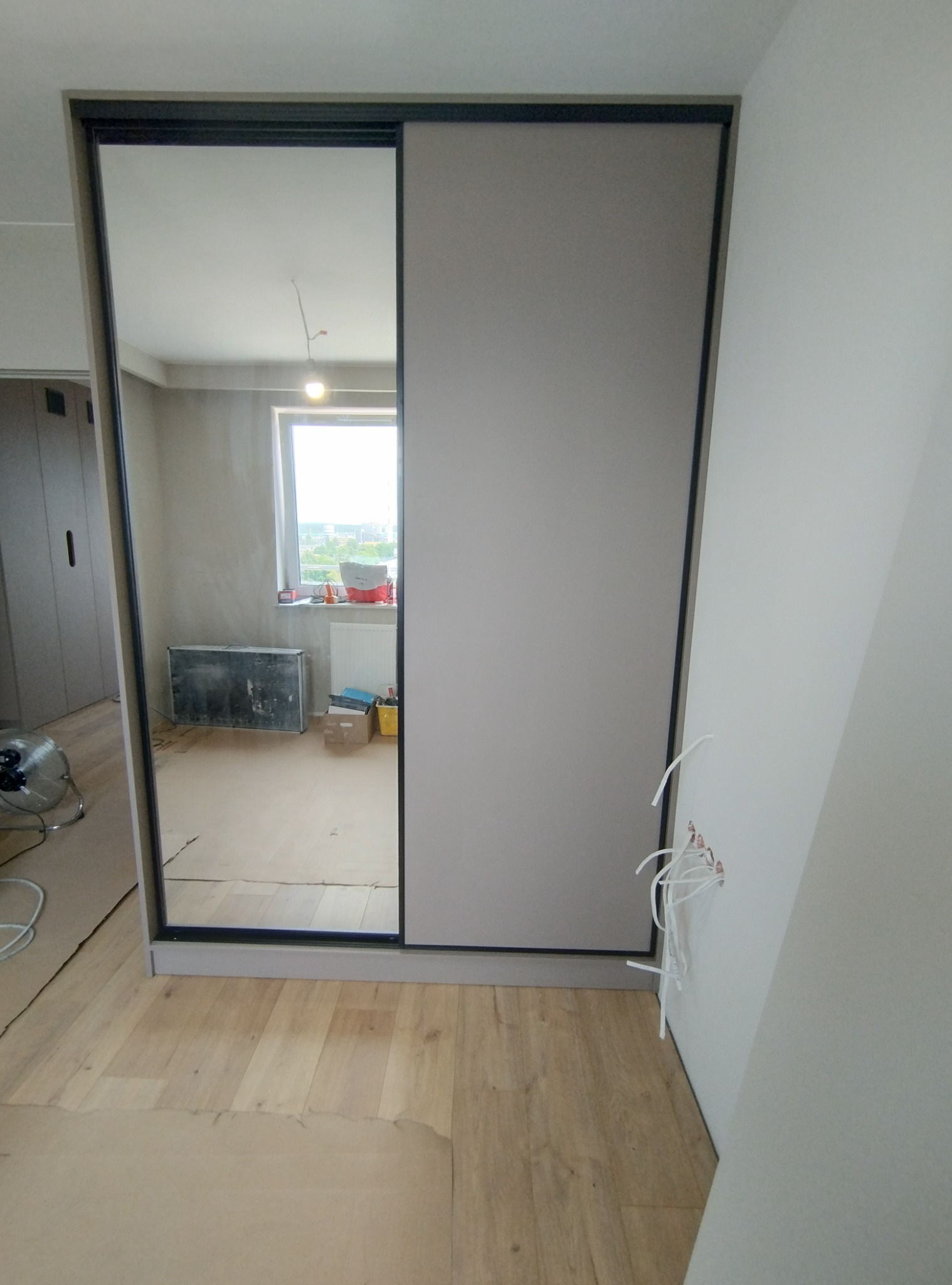 Wnętrze pokoju z zamontowaną szafą przesuwną z lustrem, widoczne wystające przewody elektryczne ze ściany, przygotowanie do wykończenia.