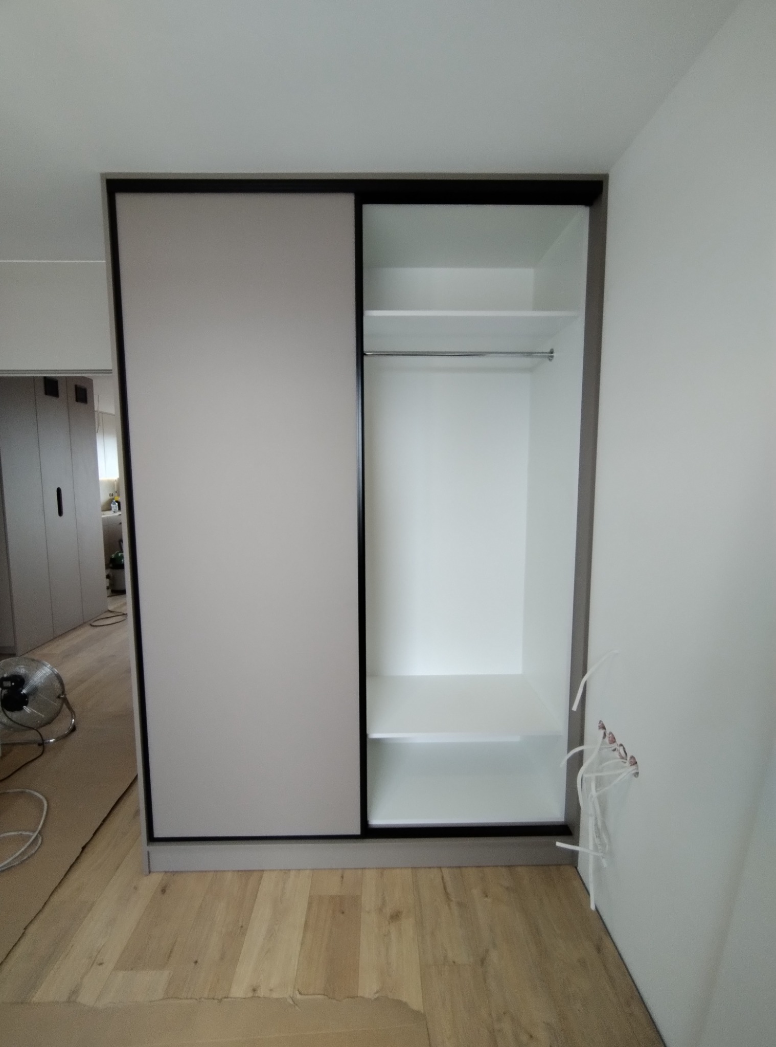 Szafa na wymiar z przesuwnymi drzwiami w kolorze szarym, z widocznym wnętrzem z półkami i drążkiem, podczas montażu, z widocznymi przewodami elektrycznymi wystającymi ze ściany.