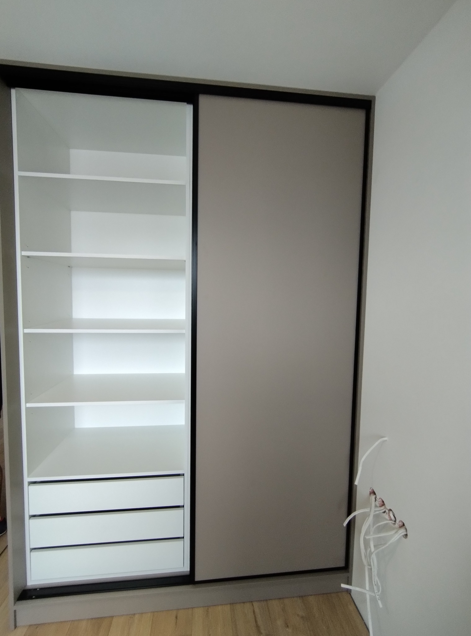 Nowoczesna szafa przesuwna z białymi półkami i szufladami oraz szarą płytą drzwiową, widoczne przewody elektryczne w ścianie.
