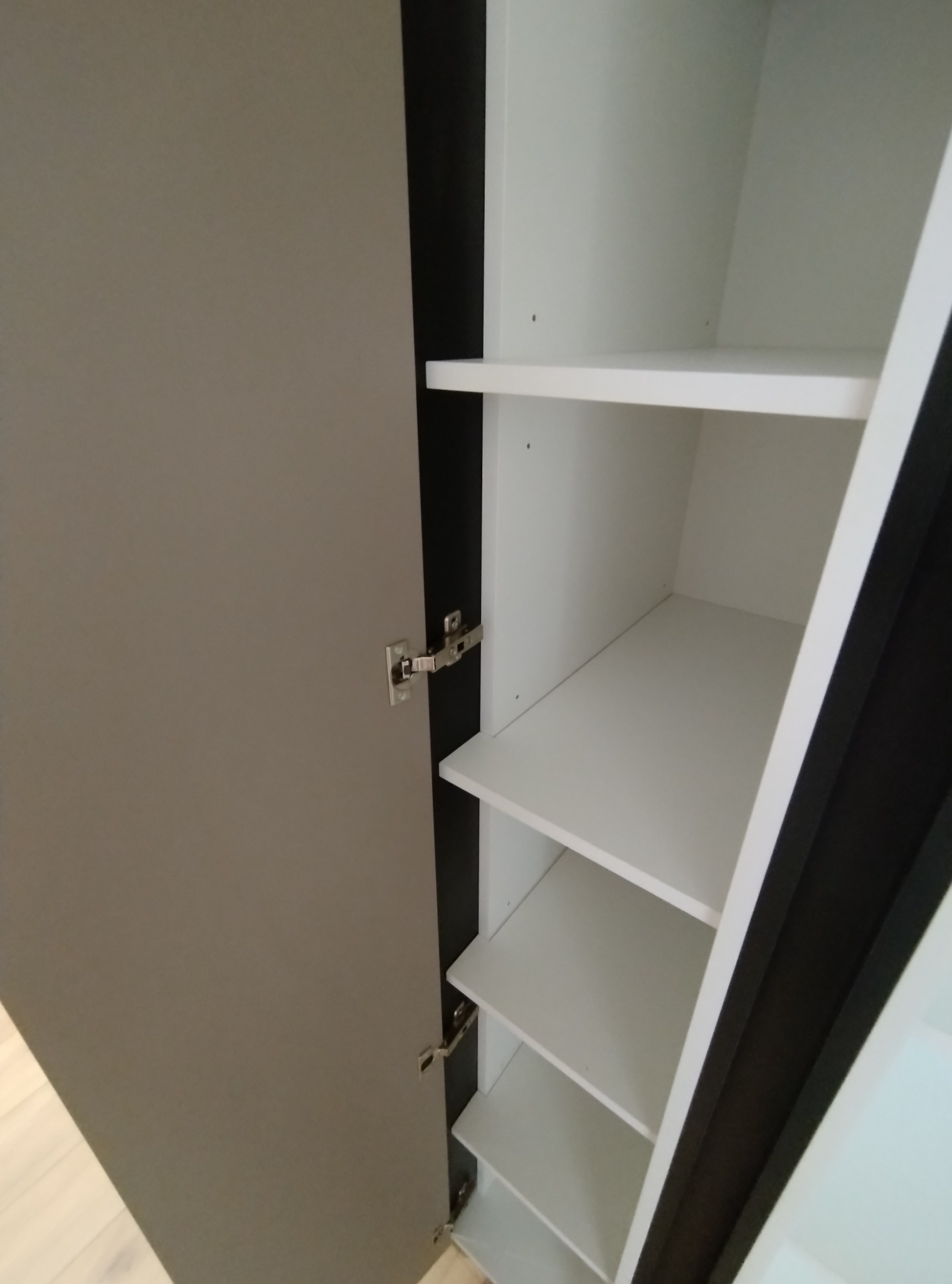 Otwarta szafa z szarymi drzwiami i białymi półkami, widoczne zawiasy.