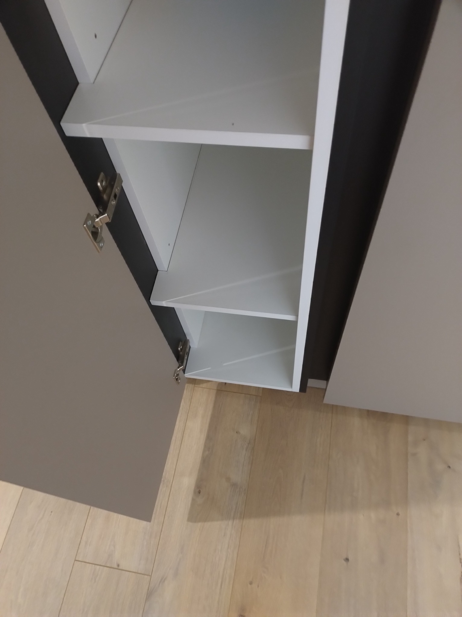 Otwarta szafka z półkami w odcieniach szarości i bieli, widoczne zawiasy, na podłodze panele w Białymstoku.