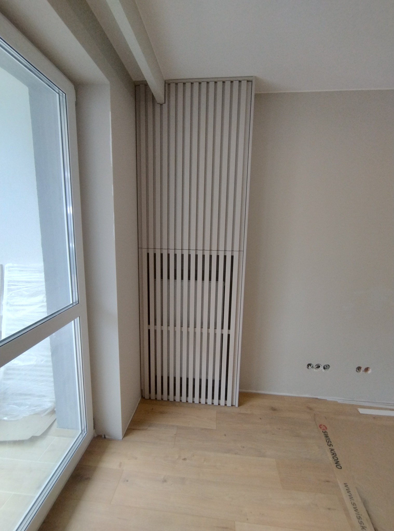 Wykończona ściana z drewnianymi lamelami w jasnym kolorze, częściowo zakrywająca grzejnik, widoczna obok okna w nowym mieszkaniu z podłogą wyłożoną panelami.