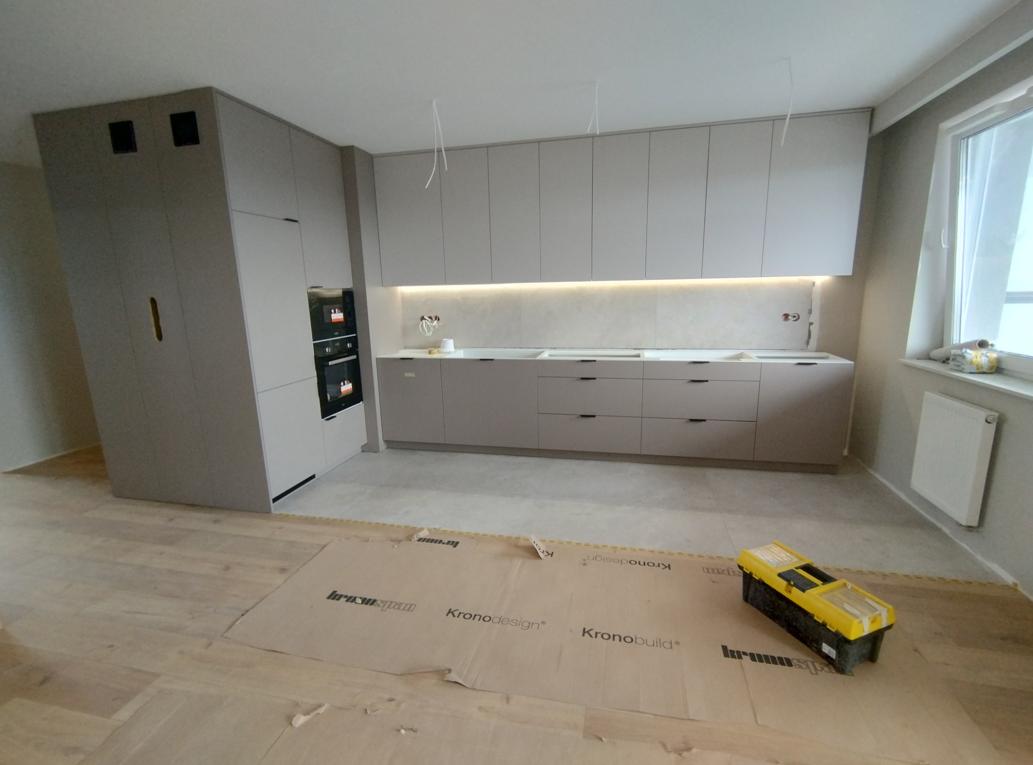 Nowoczesna, matowa kuchnia w odcieniach szarości z oświetleniem podszafkowym, widoczna skrzynka z narzędziami na podłodze zabezpieczonej kartonem.