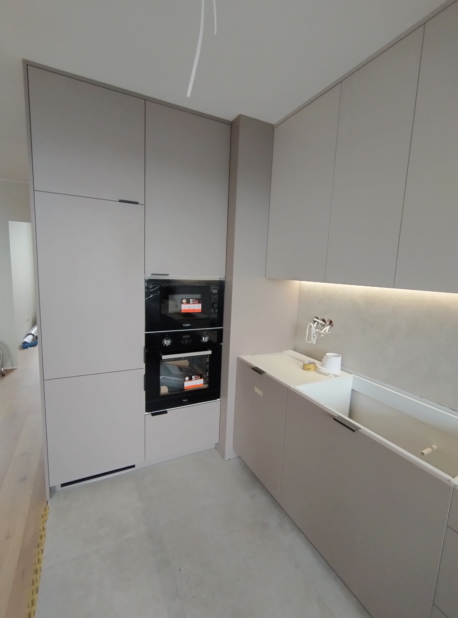 Jasnoszara zabudowa kuchenna z wbudowanym piekarnikiem i mikrofalówką, widoczne niedokończone podłączenia instalacji nad blatem.