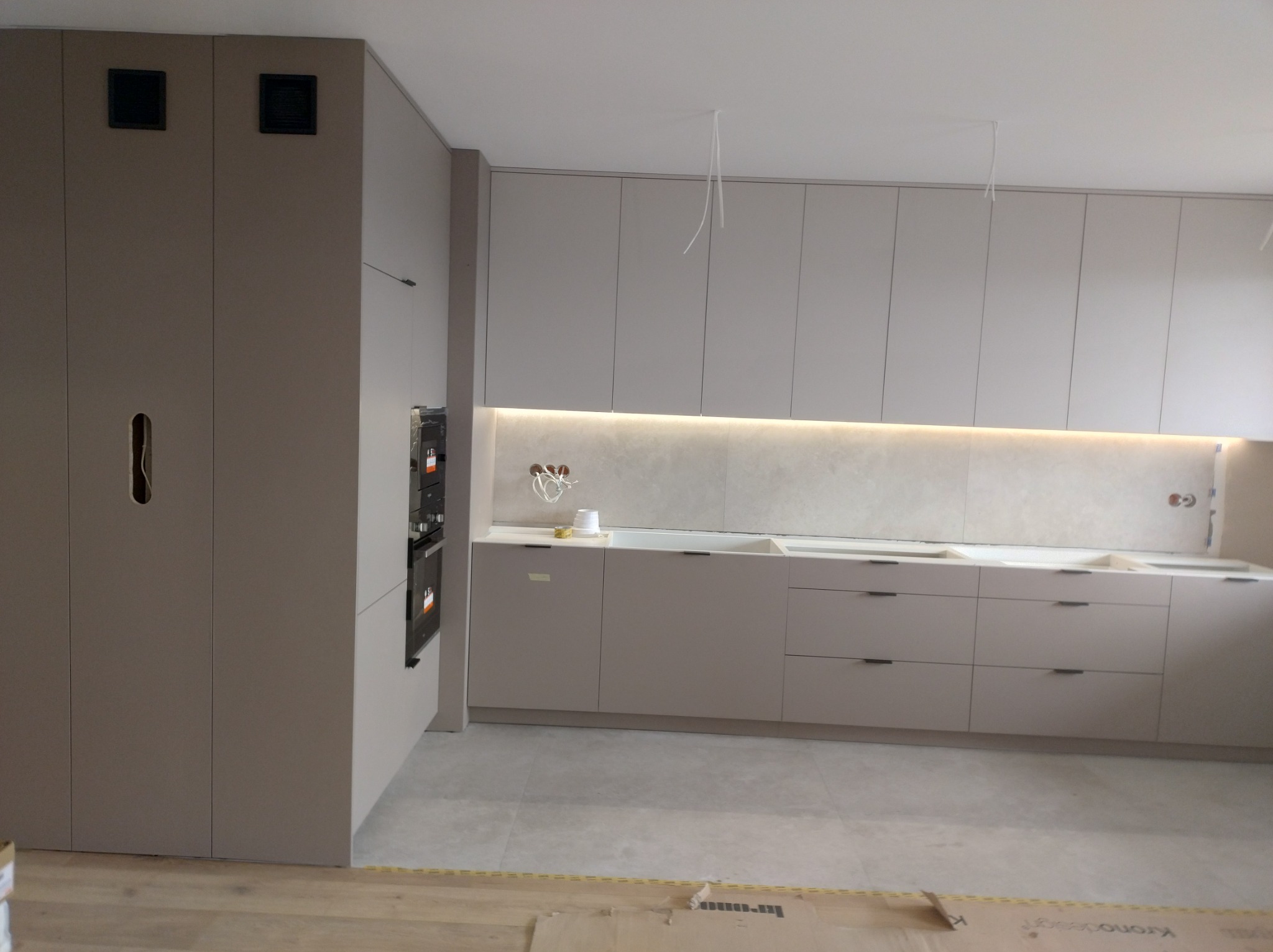 Szare, minimalistyczne meble kuchenne w trakcie montażu, widoczne niedociągnięcia wykończeniowe i brakujące elementy dekoracyjne, oświetlenie LED pod szafkami, kartony na podłodze.