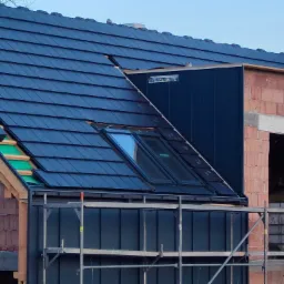 Budowa domu: fragment dachu z ciemną dach&oacute;wką ceramiczną i oknami dachowymi, widoczna konstrukcja drewniana, rusztowanie i fragment muru z cegły.