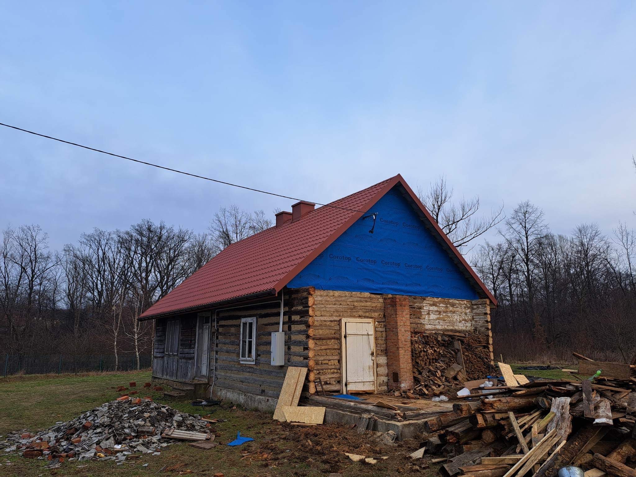 Stary drewniany dom z nowym czerwonym dachem i niebieską membraną dachową, stos drewna obok wejścia, gruz przed domem.