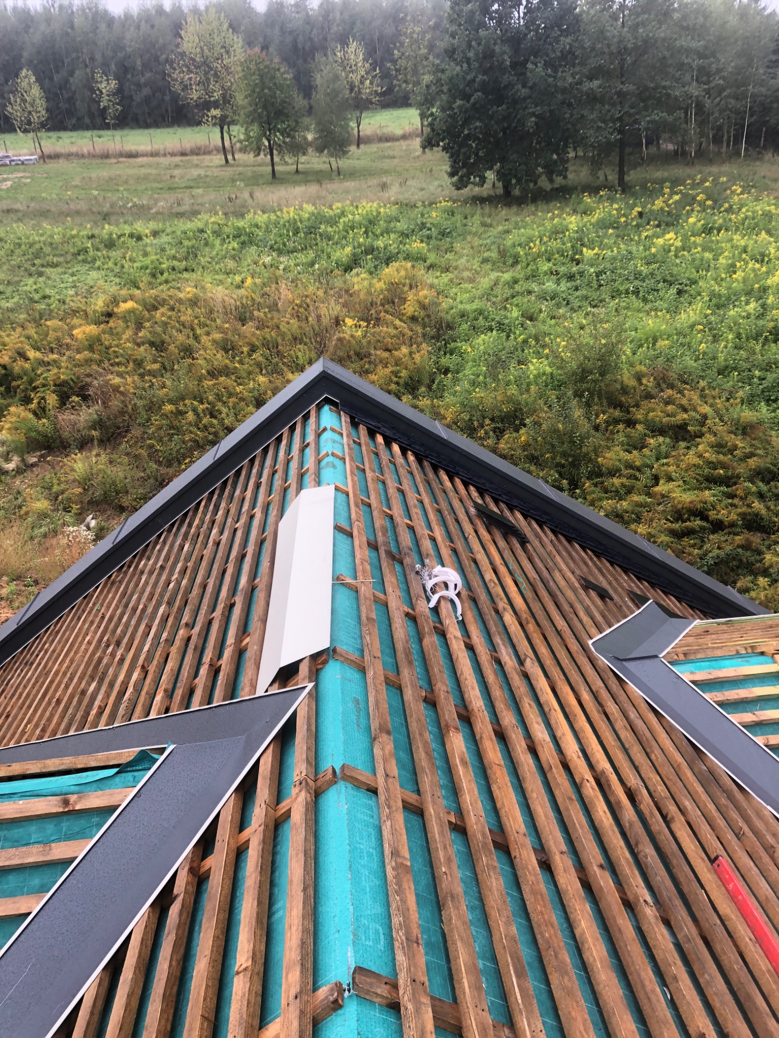 Konstrukcja dachu z drewnianymi łatami i turkusową membraną, widok z góry na tle zieleni.