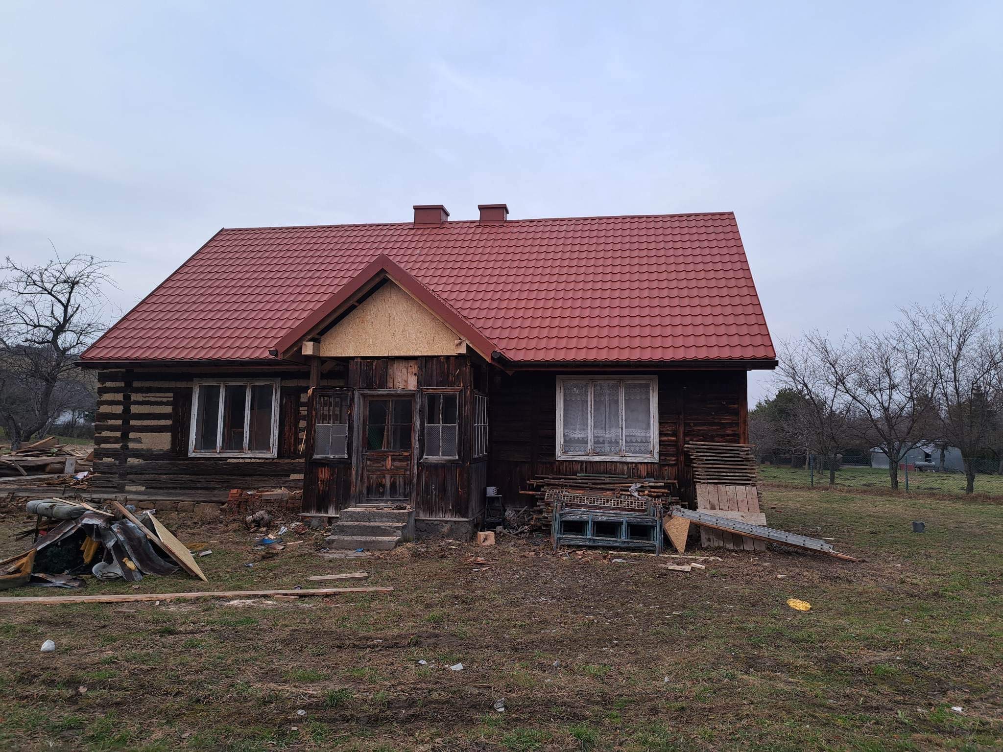 Częściowo wyremontowany, drewniany dom z nowym, czerwonym dachem z blachodachówki, widoczne elementy starej konstrukcji i materiały budowlane wokół budynku.