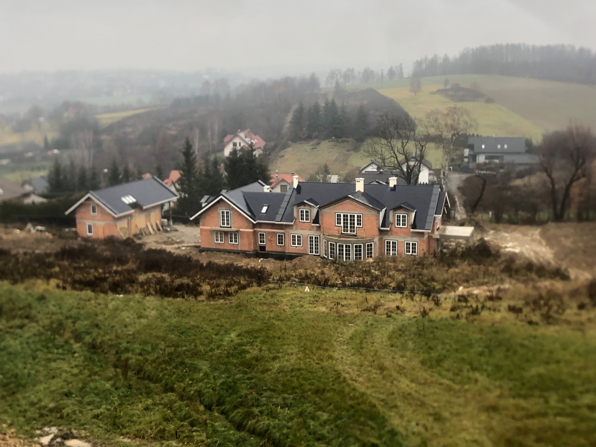 Niedokończony dom murowany z ciemną dachówką na wzgórzu w pochmurny dzień, widoczne rusztowania i drugi budynek w stanie surowym obok.