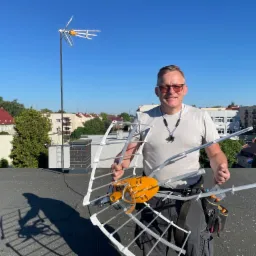 Instalator na dachu trzyma nową antenę Yagi-Uda z pomarańczowym symetryzatorem, w tle budynki mieszkalne i antena zamontowana na maszcie.