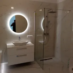 Wykończona łazienka z kabiną prysznicową typu walk-in, umywalką nablatową w kształcie łódki i okrągłym lustrem z podświetleniem LED, marmurowe płytki na ścianach i podłodze z widocznym odbiciem...