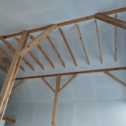 BWG- Big Work Group - Drewniana konstrukcja szkieletowa dachu w surowym stanie, widoczna wewnątrz jasnego pomieszczenia z oknem.