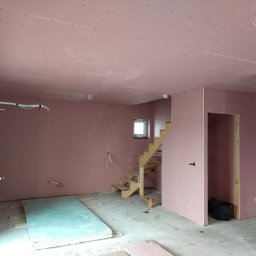 BWG- Big Work Group - Surowe wnętrze z płyt gipsowo-kartonowych w odcieniach różu, widoczne drewniane schody, okno, instalacje elektryczne i stos płyt na podłodze.